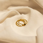 The Black Abyss Ring Set in 18K Gold features three delicate gold rings with slim bands and a small black gemstone, displayed on soft, cream-colored fabric.