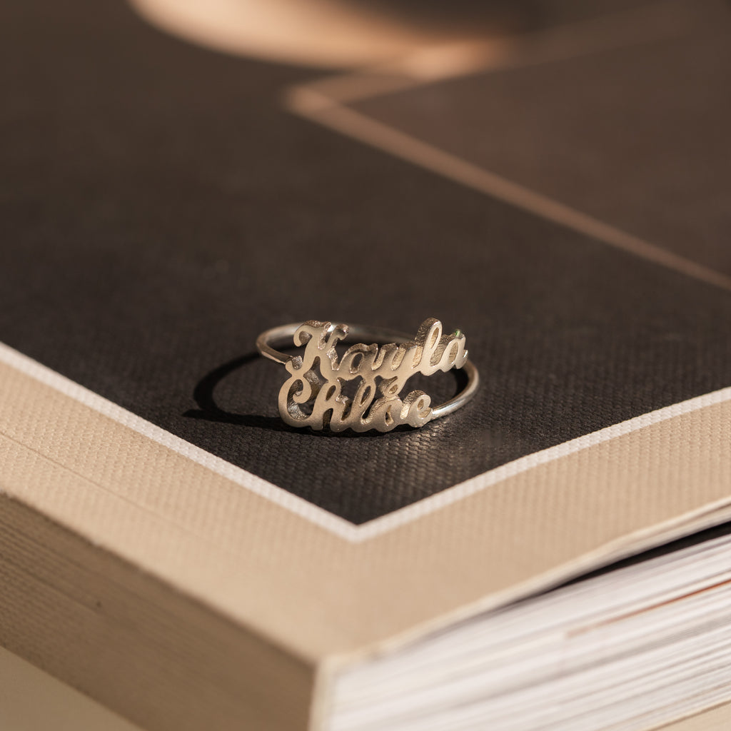 The London Duo Name Ring, engraved with Kayla and Chloe in silver, rests elegantly on the corner of a closed book.