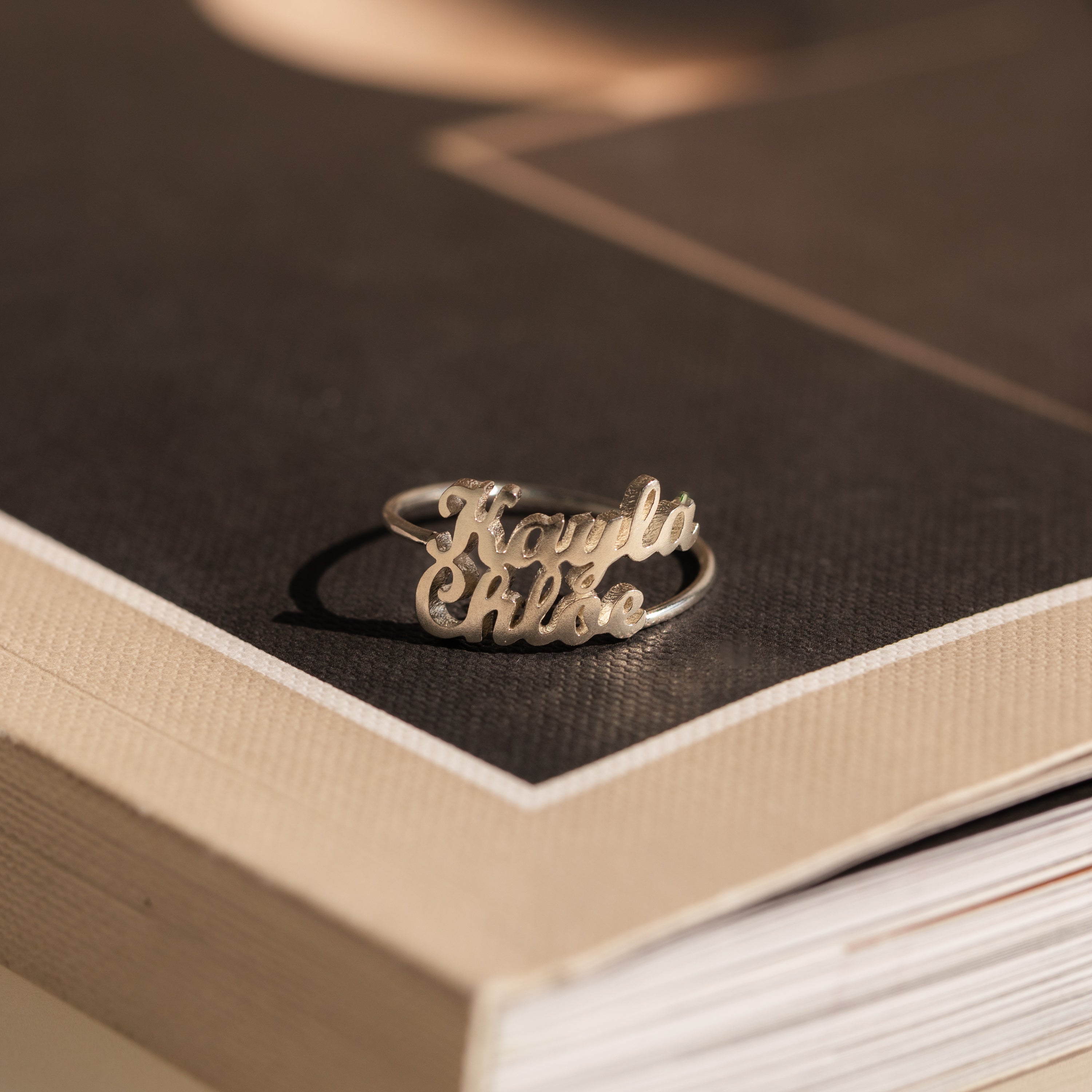 The London Duo Name Ring, engraved with Kayla and Chloe in silver, rests elegantly on the corner of a closed book.