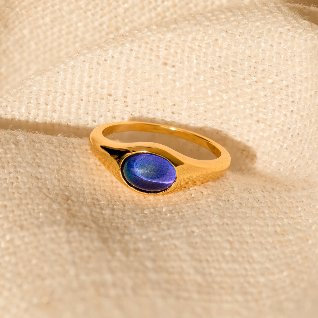 The Bold Mood Ring, featuring a blue oval stone and a sculptural gold design, rests on textured cream fabric, its elegant allure heightened by the way it catches the light.