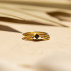 A gold dainty ring band with a black gemstone and small clear stones rests on a light surface in soft sunlight—a stunning piece from the Black Abyss Ring Set in 18K Gold.