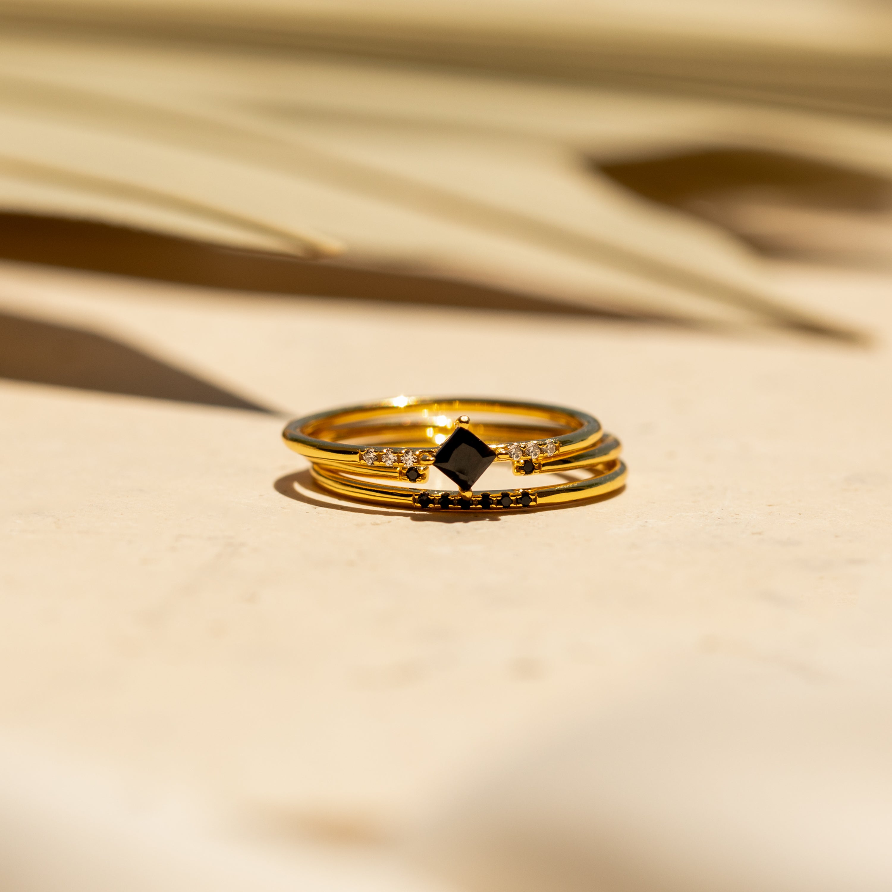 A gold dainty ring band with a black gemstone and small clear stones rests on a light surface in soft sunlight—a stunning piece from the Black Abyss Ring Set in 18K Gold.