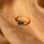 The Bold Mood Ring, featuring a dark iridescent stone and sculptural silhouette, is displayed on soft tan fabric.