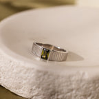 The Olive Peridot Signet Ring features vertical grooves and a rectangular green gemstone, offering bold elegance to any look. Shown on a white textured surface.
