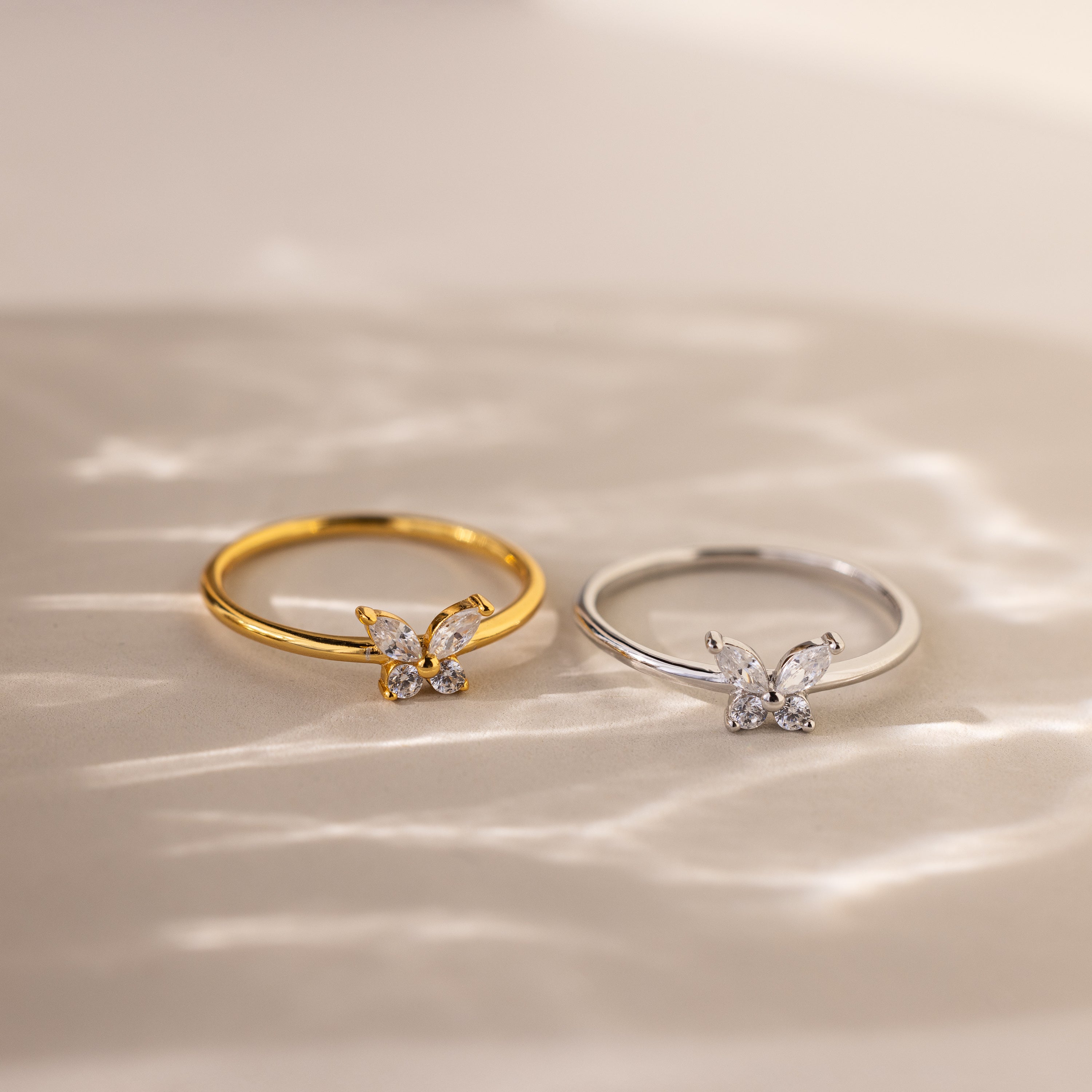 The Mariposa Diamond Ring, with gold and silver butterfly-shaped CZ settings, shines on a reflective surface—ideal as a butterfly promise ring or friendship ring.