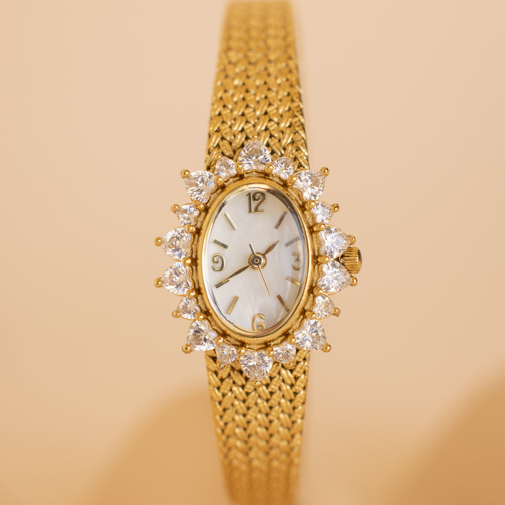 The Pave Heart Timepiece features a diamond-trimmed oval face and a woven gold band, elegantly displayed on a beige background.
