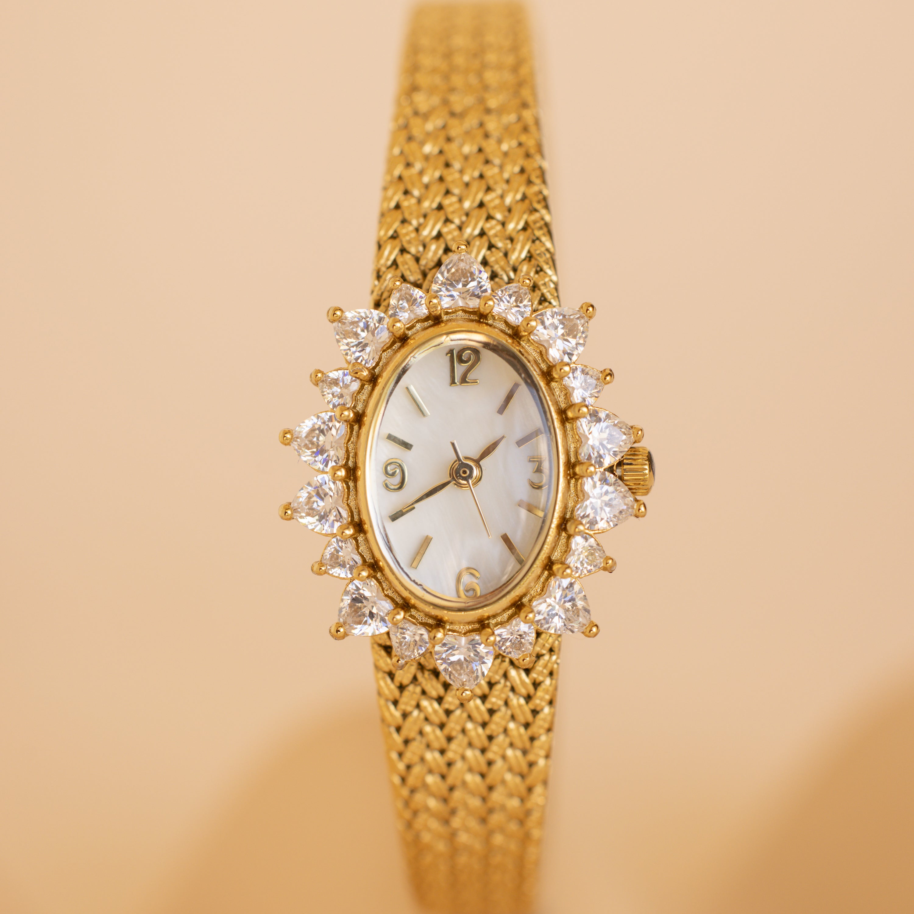 The Pave Heart Timepiece features a diamond-trimmed oval face and a woven gold band, elegantly displayed on a beige background.