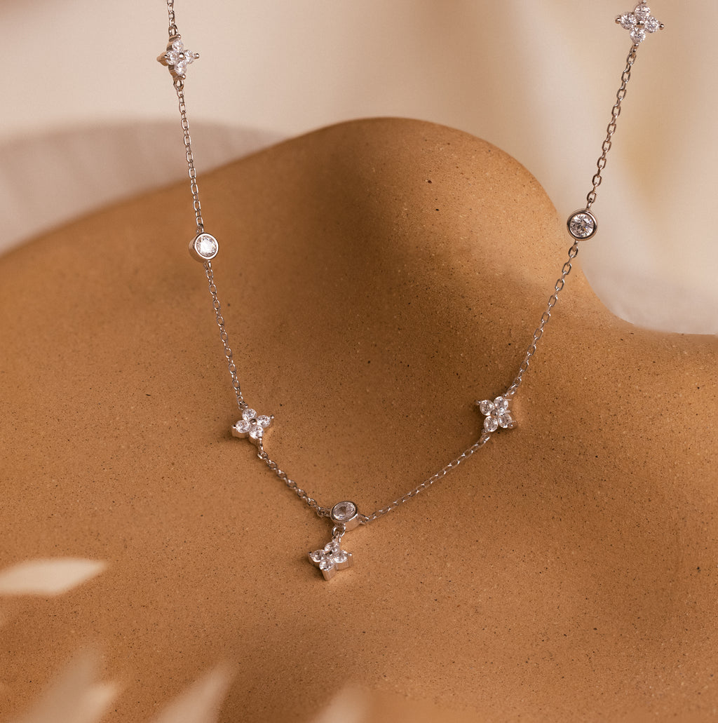 The Ivy Diamond Station Necklace features small crystals, flower-shaped charms, and diamond accents, elegantly displayed on a curved tan surface.