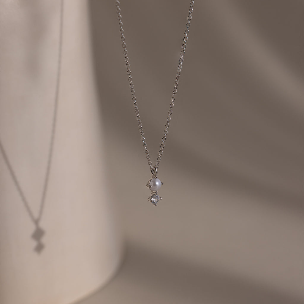 The Pearl Diamond Drop Necklace in Silver features a small pearl and diamond pendant, casting a delicate shadow on beige—ideal bridal jewelry.