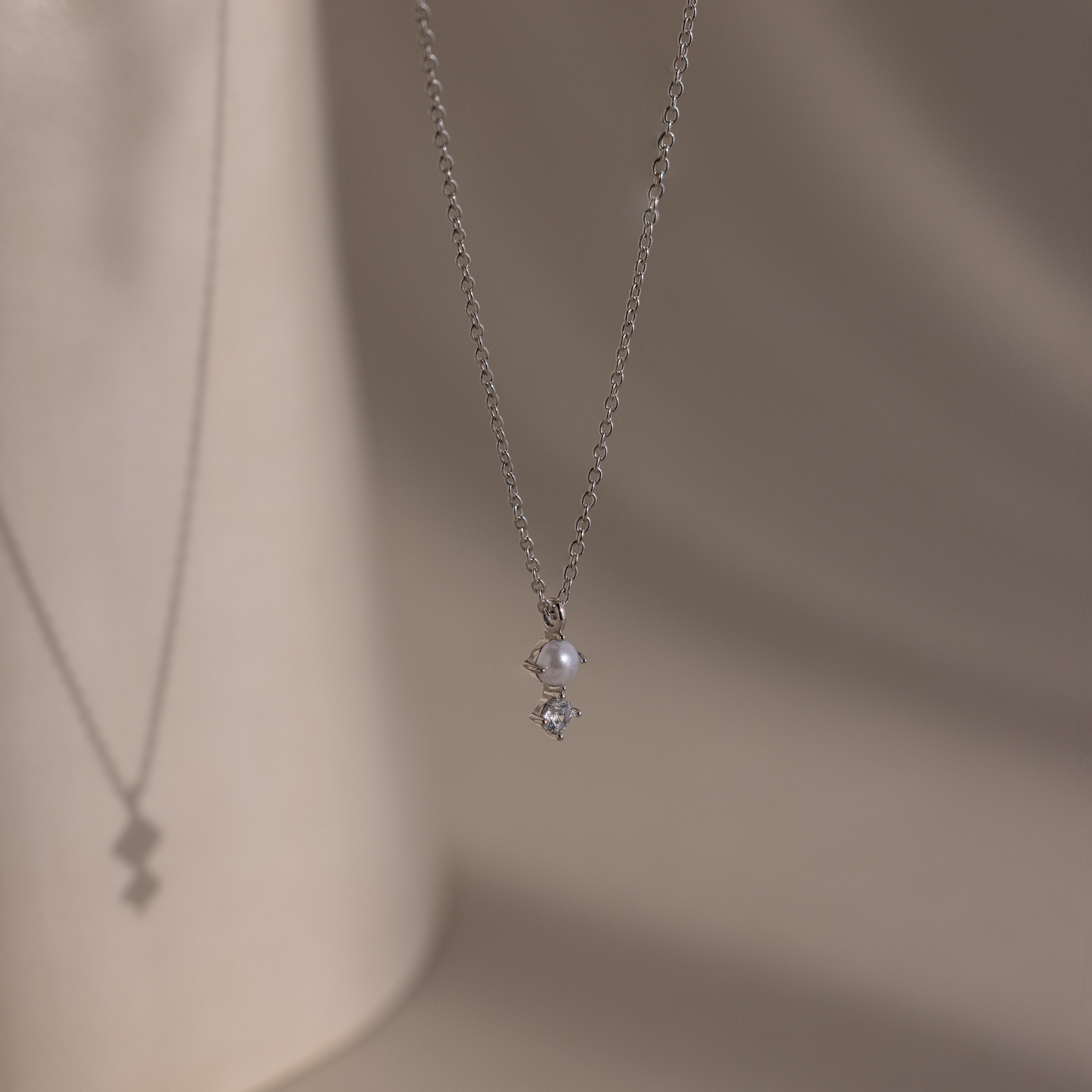 The Pearl Diamond Drop Necklace in Silver features a small pearl and diamond pendant, casting a delicate shadow on beige—ideal bridal jewelry.