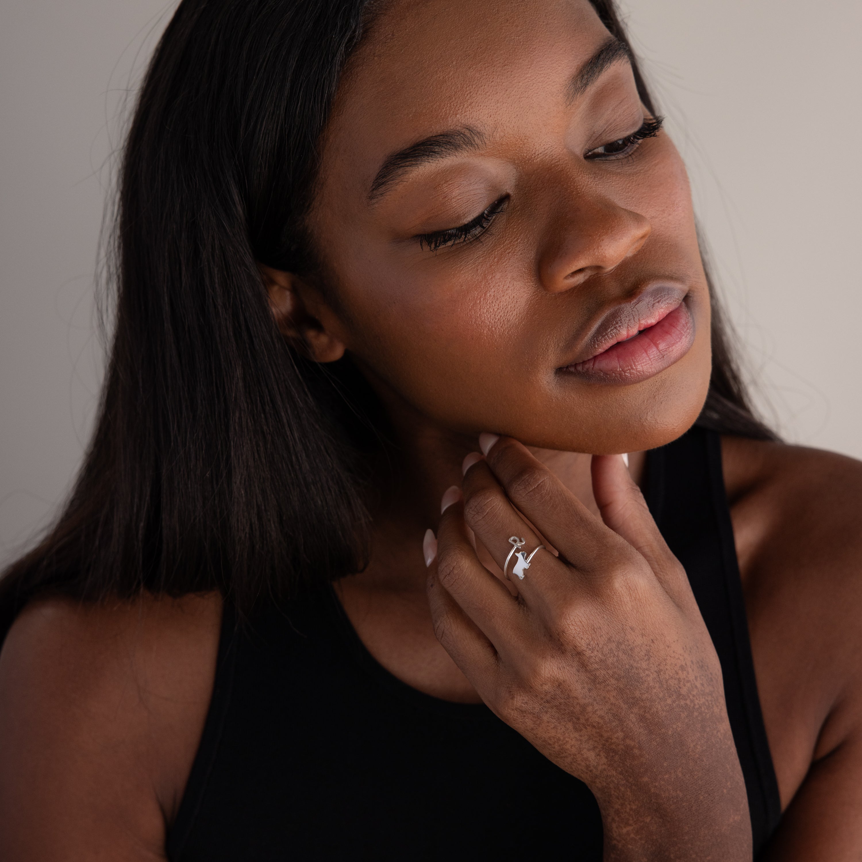 A woman with long hair, dressed in a black top, gently touches her face while showcasing the Duo Pet Initial Wrap Ring in Sterling Silver on her finger.