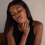 A woman with long dark hair, wearing a black tank top and looking down, touches her chin while displaying the Alex Dainty Letter Ring in Sterling Silver—a personalized piece that adds subtle elegance to her look.