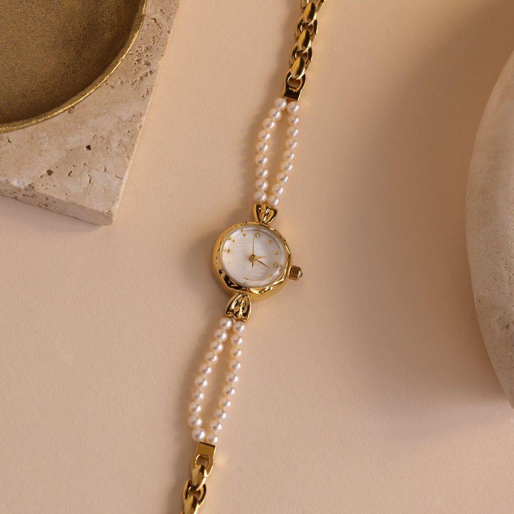 The Beaded Pearl Timepiece in 18K Gold is displayed on a beige surface with stone accents nearby.