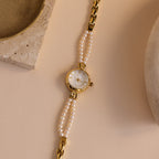 The Beaded Pearl Timepiece in 18K Gold is displayed on a beige surface with stone accents nearby.