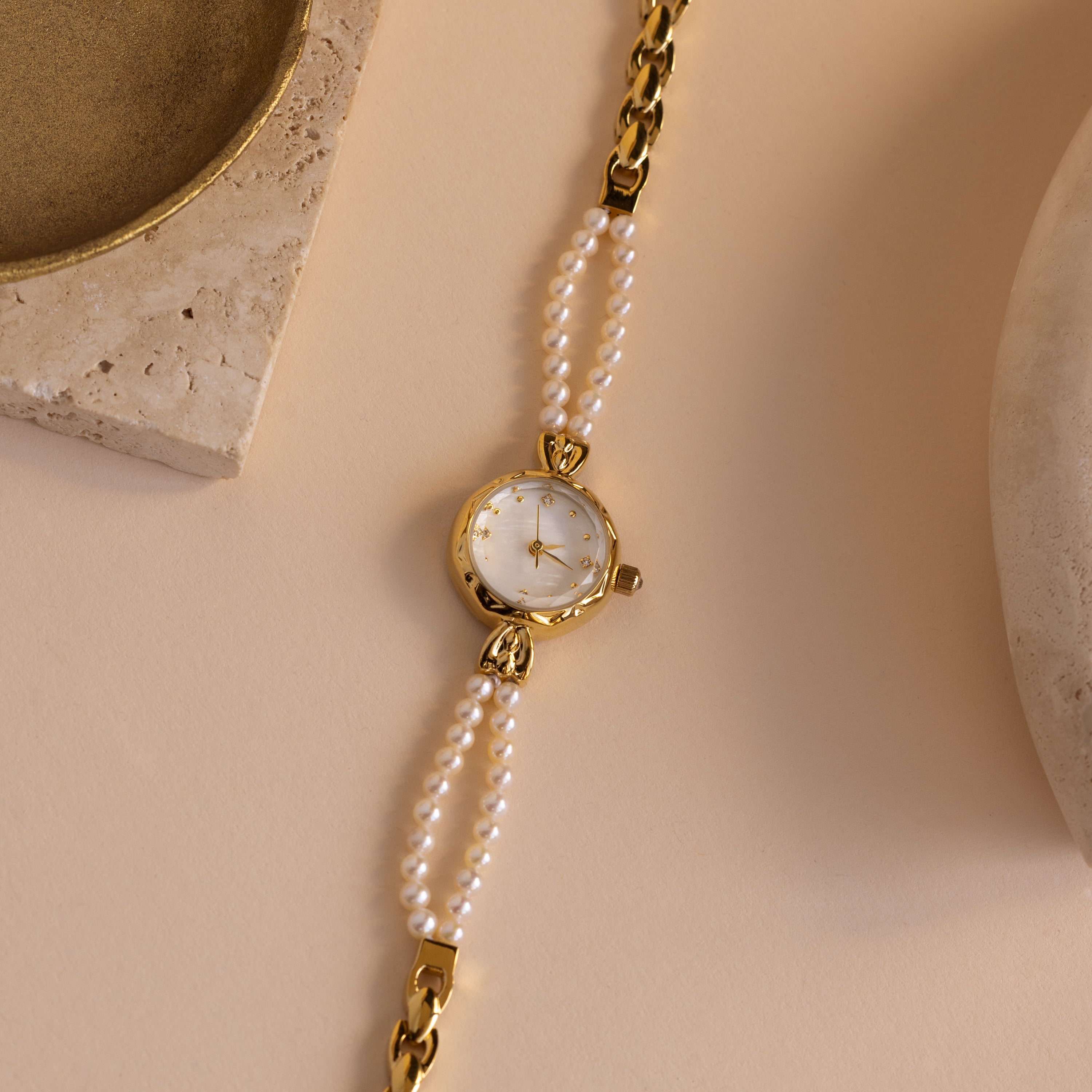 The Beaded Pearl Timepiece in 18K Gold is displayed on a beige surface with stone accents nearby.