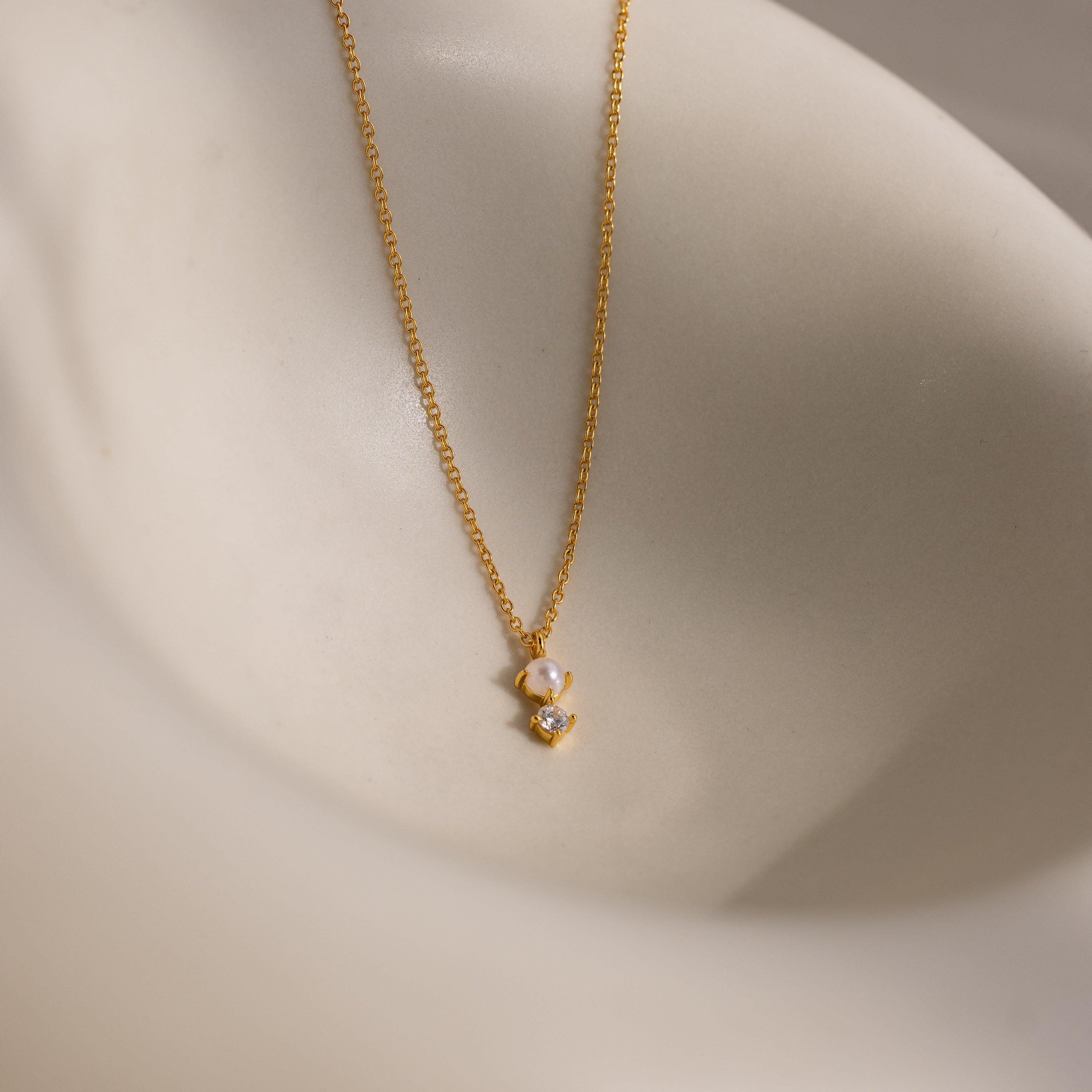 The Pearl Diamond Drop Necklace features a gold chain with a delicate pearl and diamond pendant, elegantly displayed on a cream surface. Ideal as a bridal necklace or for special occasions.
