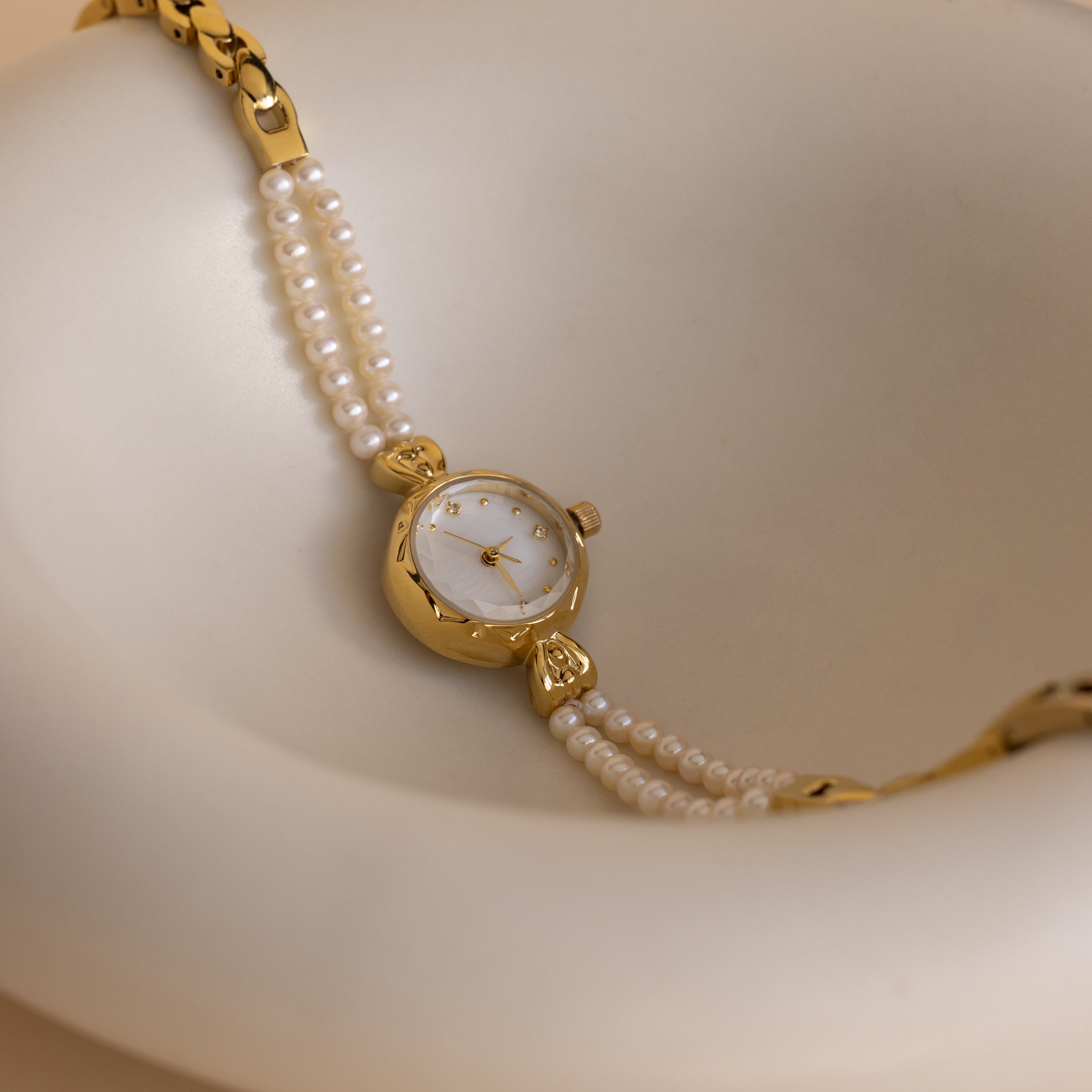 Beaded Pearl Timepiece in 18K Gold displayed on a smooth, cream-colored curved surface.