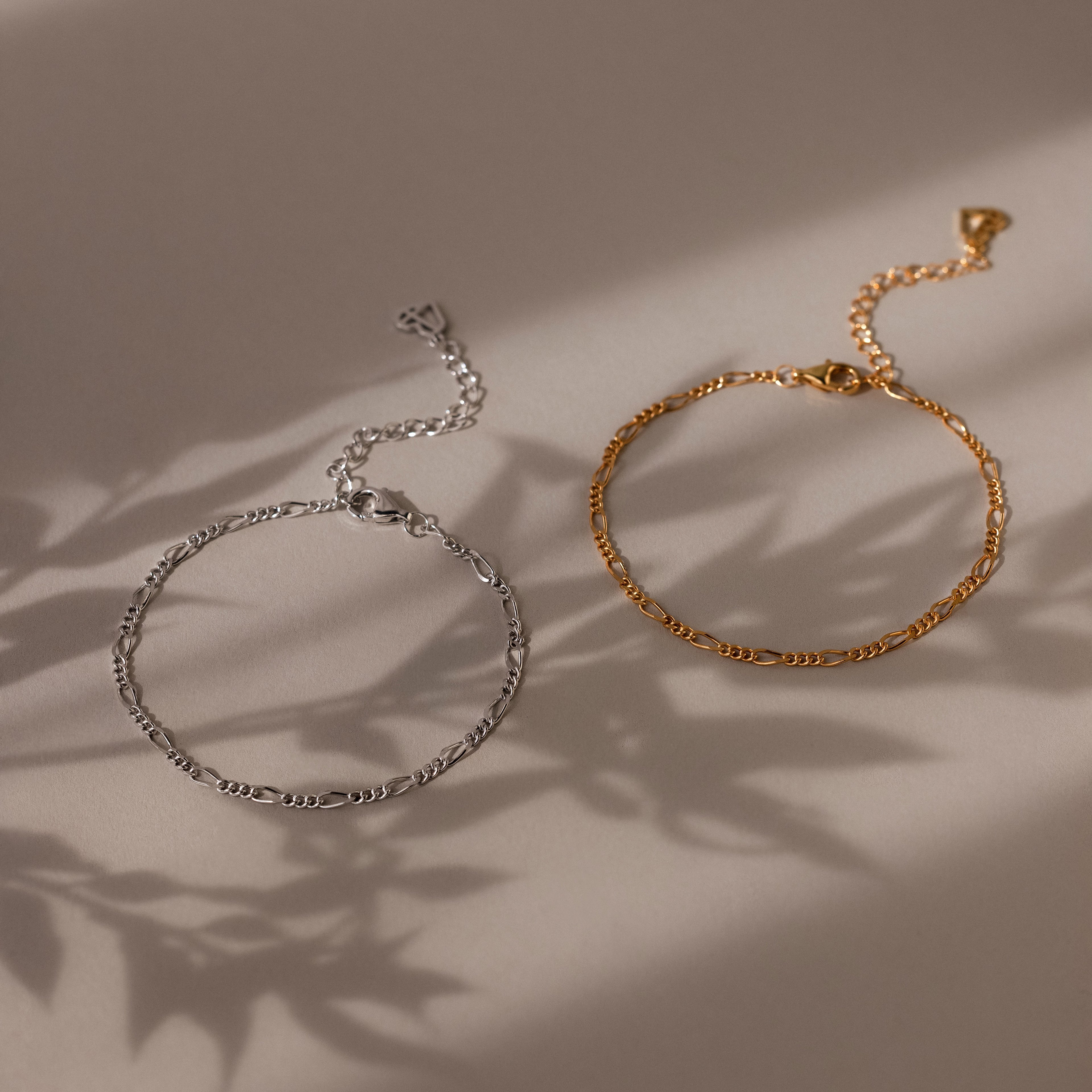 Two chain bracelets, one silver and one gold, on a beige surface with soft shadows.