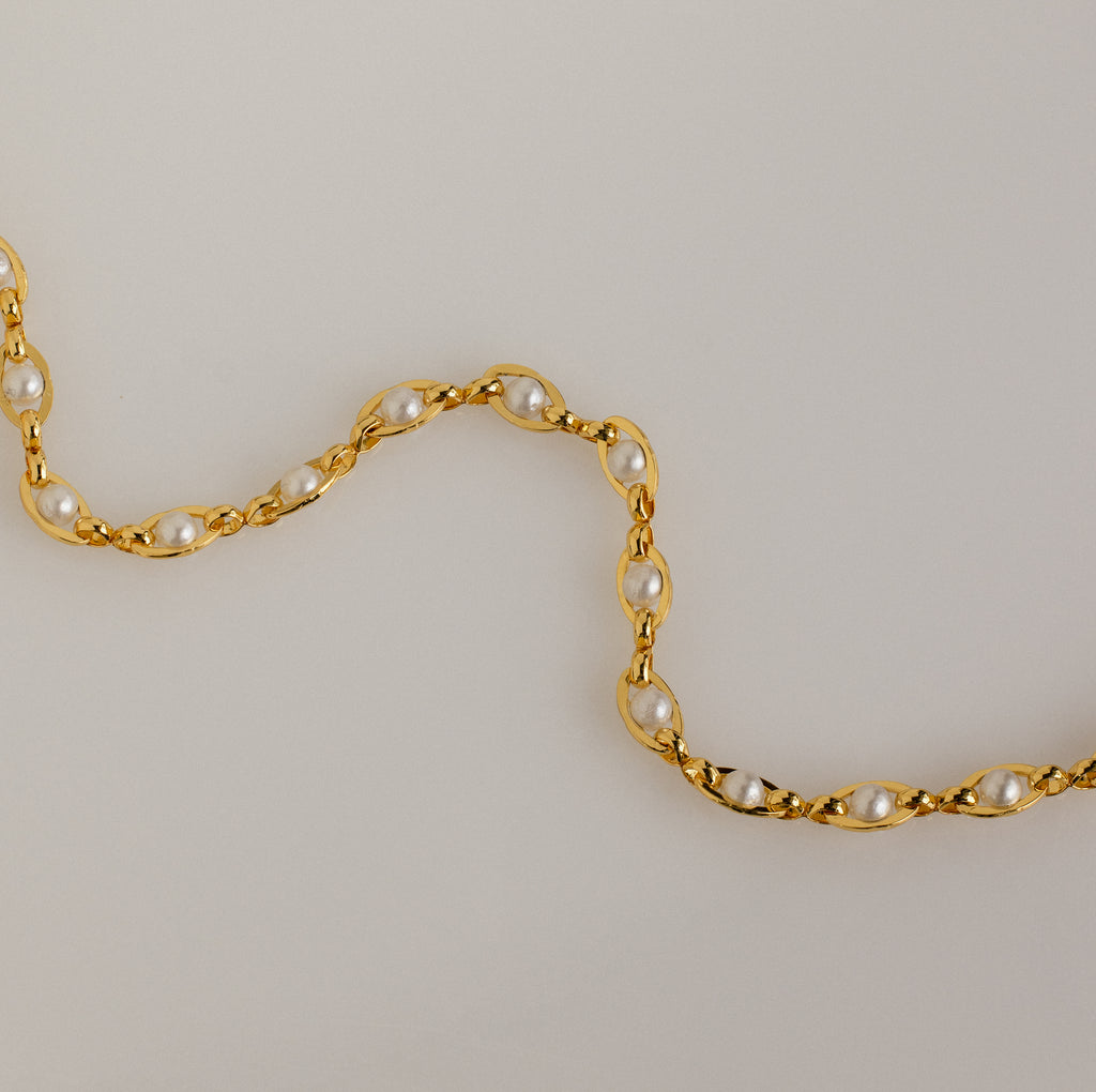 An elegant jewelry piece, the Eternity Pearl Bracelet in 18K Gold features white pearls in a wavy pattern on a light beige background.