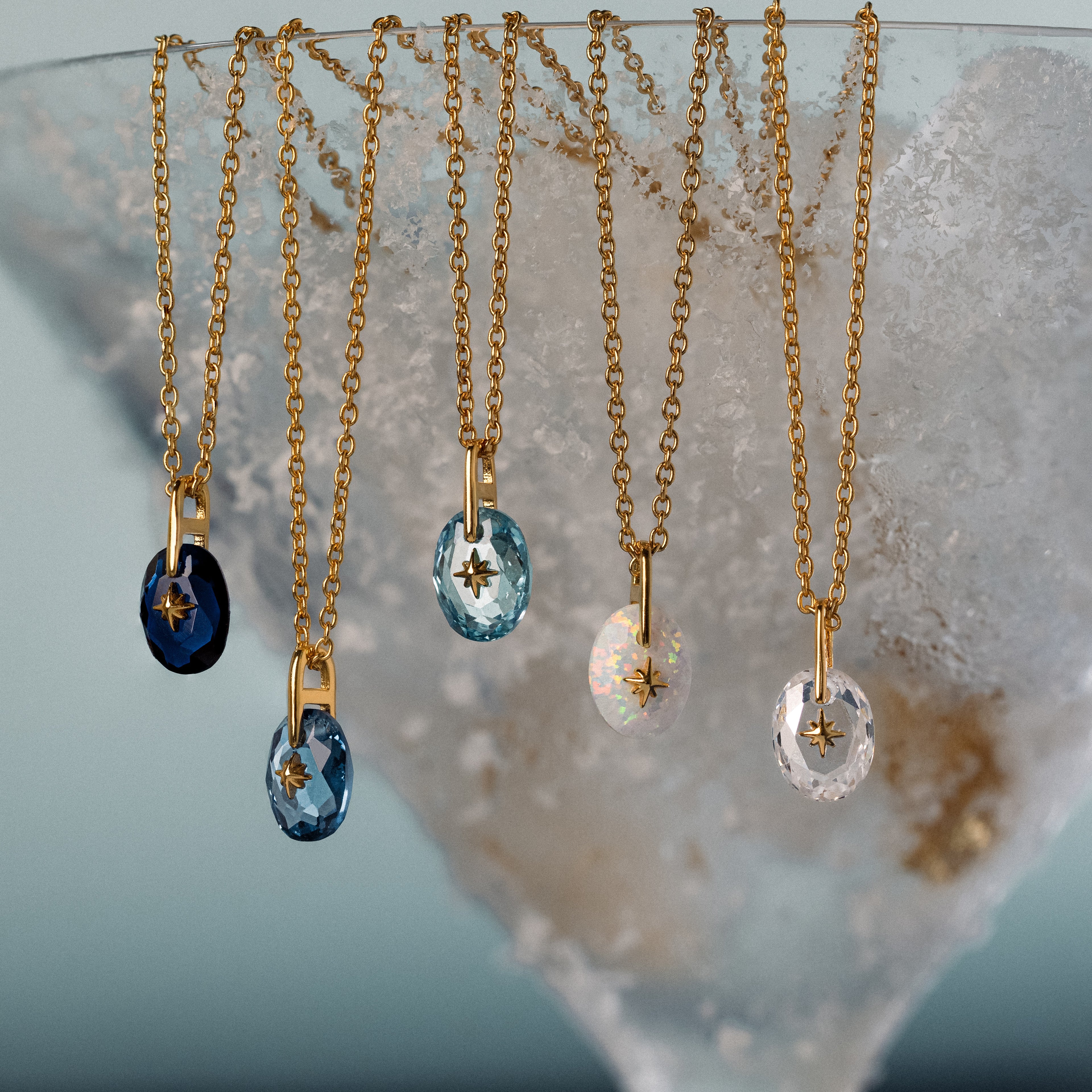 Five gold necklaces with oval gemstone pendants, including the North Star Birthstone Signet Necklace in Sapphire, elegantly hang on a textured frosted surface.