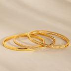Three gold bangles, including the Lace Diamond Bangle in 18K Gold with round diamonds and an open station design, are displayed on a neutral beige surface.