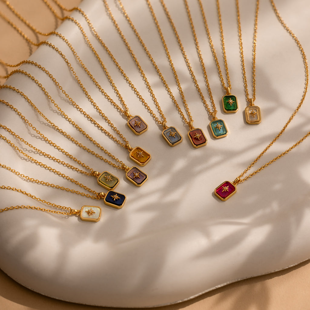 North Star Birthstone Necklaces with vibrant rectangular crystal pendants are arranged in a semi-circle on a white, wavy surface.