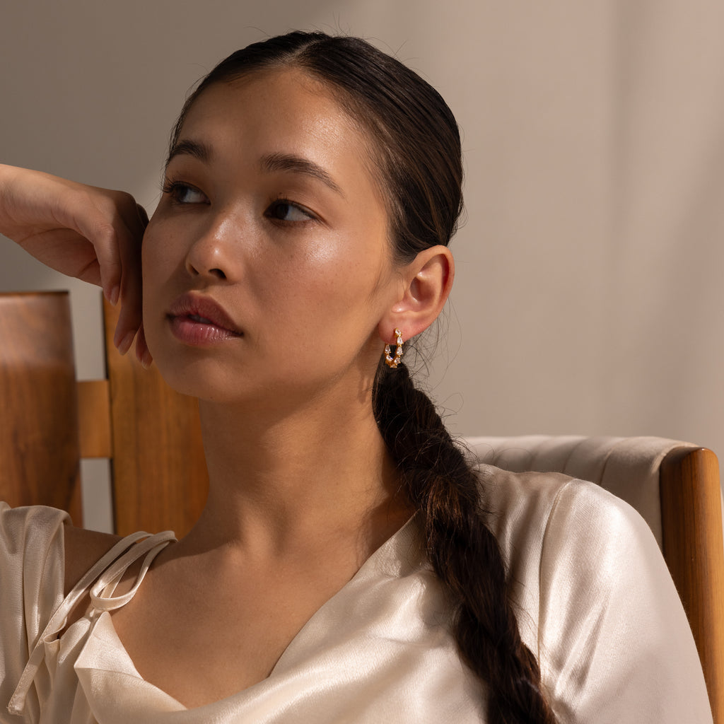 Woman with long braided hair wears Inside Out Pear Diamond Hoops and a satin top, sitting on a wooden chair and looking away.