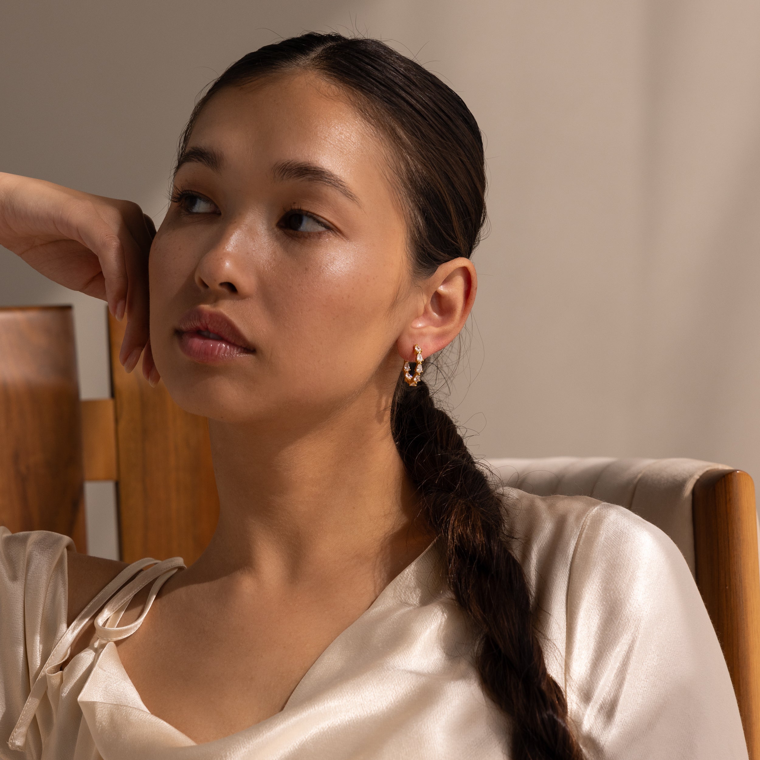 Woman with long braided hair wears Inside Out Pear Diamond Hoops and a satin top, sitting on a wooden chair and looking away.