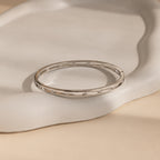 The Lace Diamond Bangle, adorned with sparkling diamonds, lies on an off-white textured surface, highlighting the refined elegance and luxurious appeal of this exquisite jewelry piece.