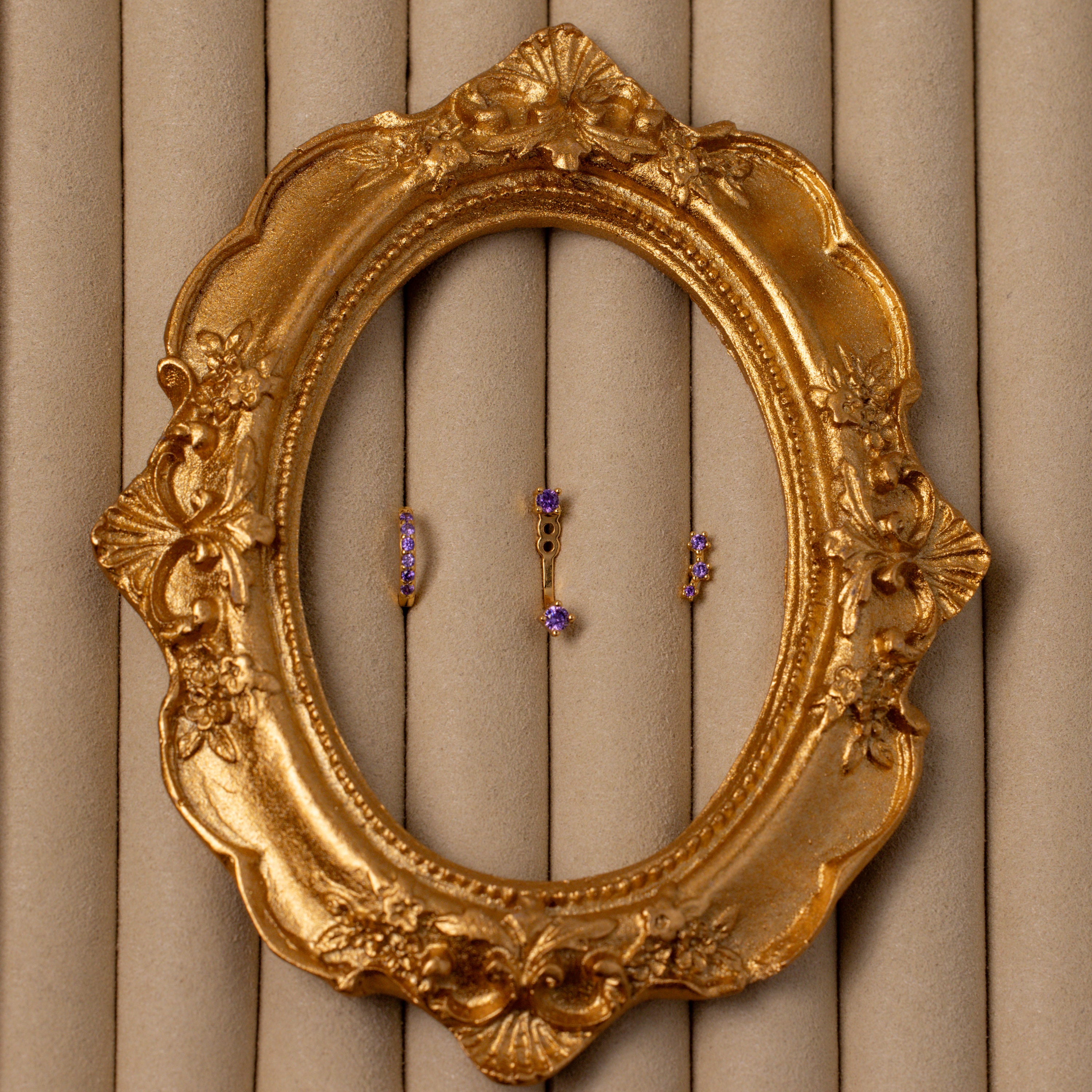 The Amethyst Earring Trio Set features three small purple birthstone earrings in a gold ornate oval frame on a beige padded background—an elegant gold set, perfect for gifting.