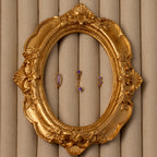 The Birthstone Earring Trio Set, featuring three small gold and purple earrings, is displayed inside an ornate oval gold frame on a beige textured background.