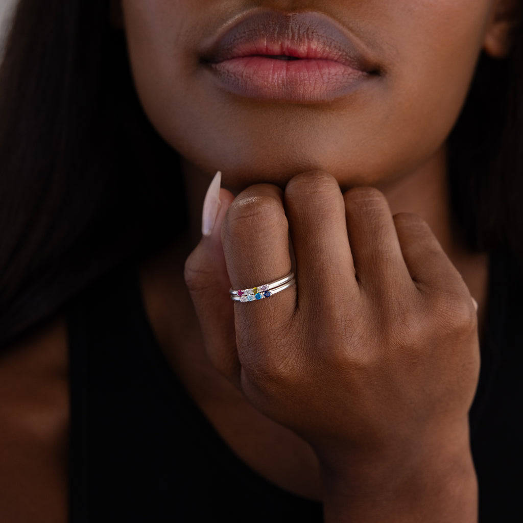 A woman rests her chin on her hand, highlighting the Mini Birthstone Ring in Sterling Silver adorned with colorful gemstones on her finger.