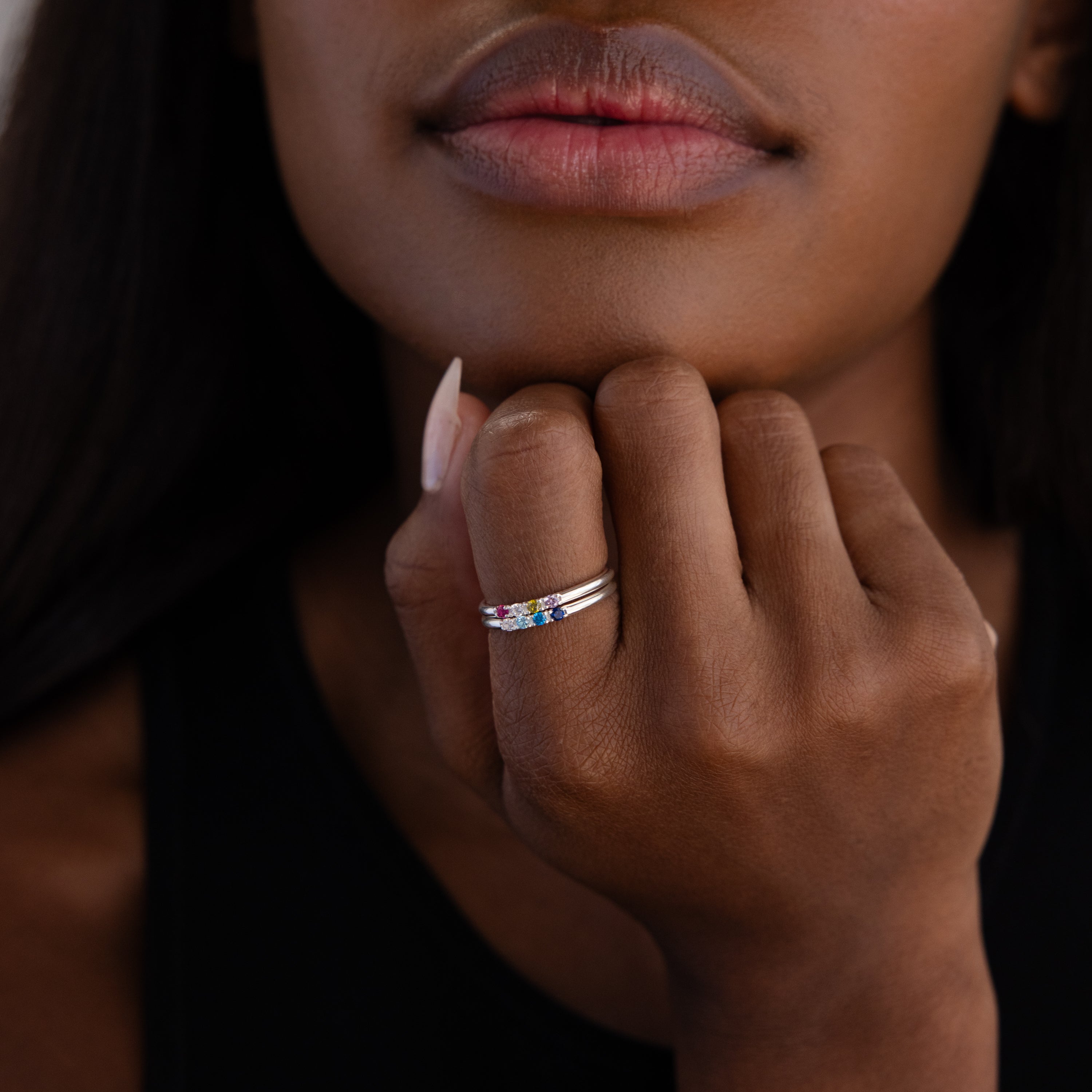 A woman rests her chin on her hand, highlighting the Mini Birthstone Ring in Sterling Silver adorned with colorful gemstones on her finger.