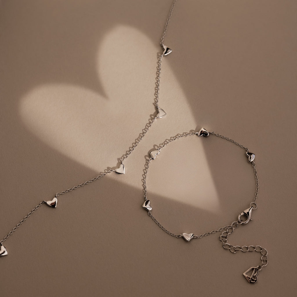 The Heart Station Bracelet in Sterling Silver features delicate heart charms and casts a soft heart-shaped shadow on a beige background—ideal as romantic jewelry or a stackable, elegant bracelet.