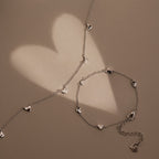 The Heart Station Necklace in Silver features delicate heart charms and rests on a beige surface, casting romantic, heart-shaped shadows in the background.