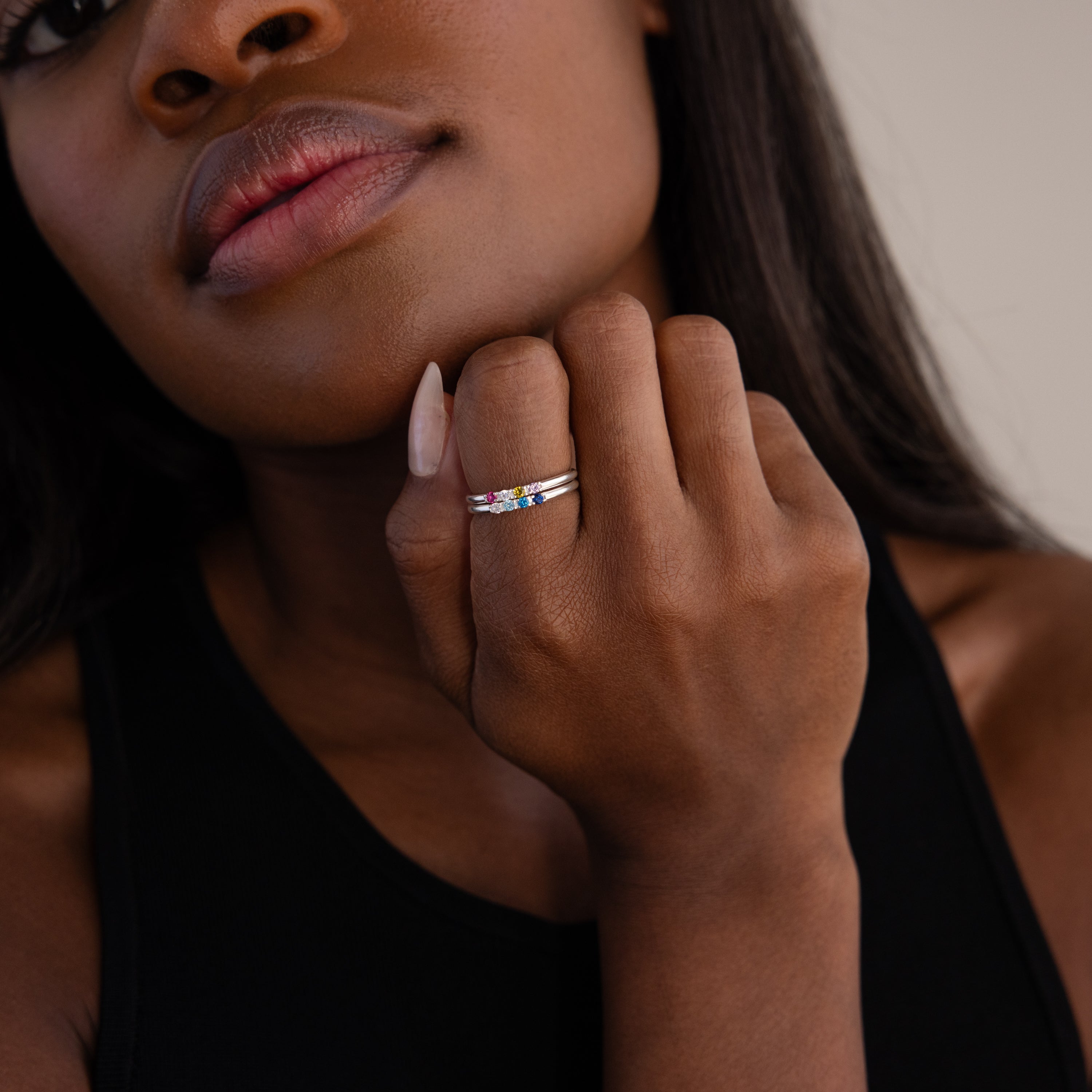 A woman in a black top showcases the Mini Birthstone Ring in Sterling Silver, featuring colorful personalized stones, on her hand near her face.
