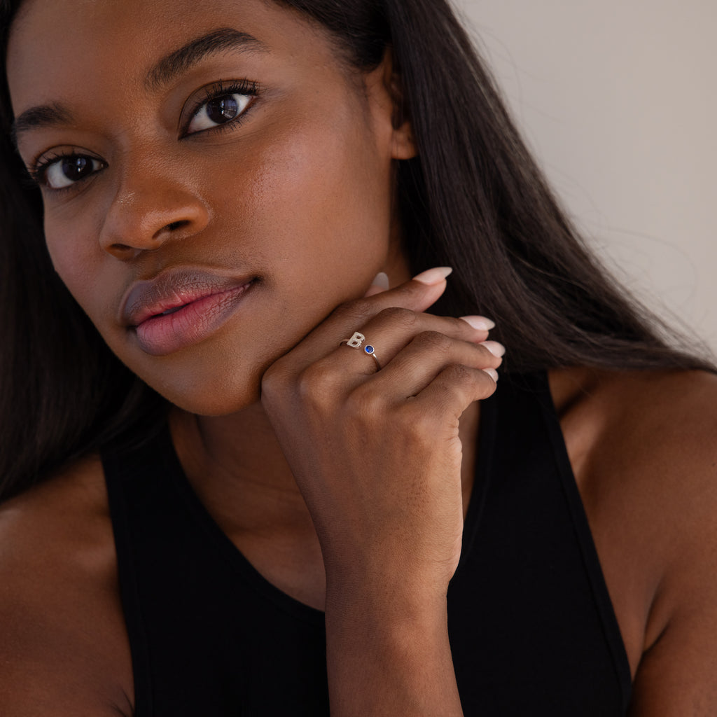 Woman with long dark hair wearing a black top displays the Alex Initial Birthstone Ring in Sterling Silver, featuring a striking blue stone on her finger.