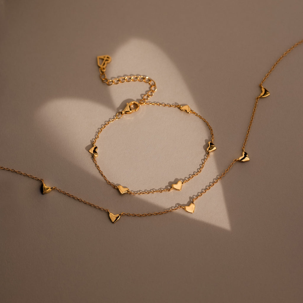 Heart Station Necklace with delicate heart charms on a cable chain, displayed on a beige surface casting a heart-shaped shadow.