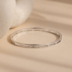 The Lace Diamond Bangle in Sterling Silver, featuring delicate round diamonds, is shown on a beige surface.