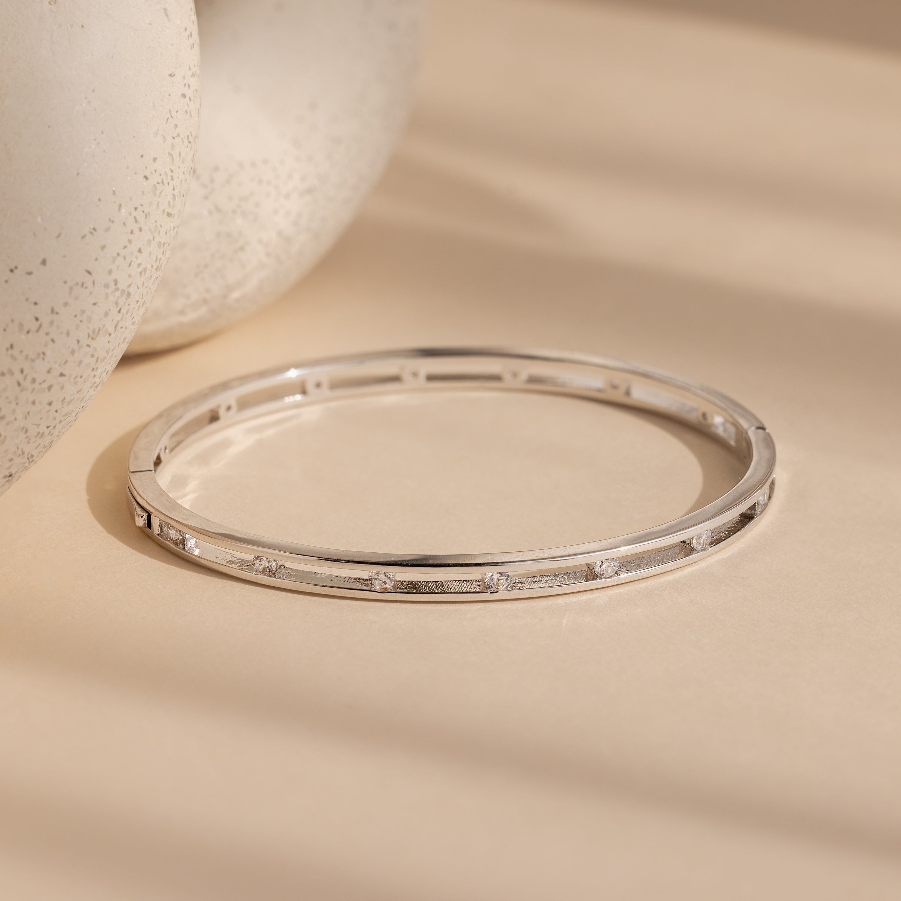 The Lace Diamond Bangle in Sterling Silver, featuring delicate round diamonds, is shown on a beige surface.