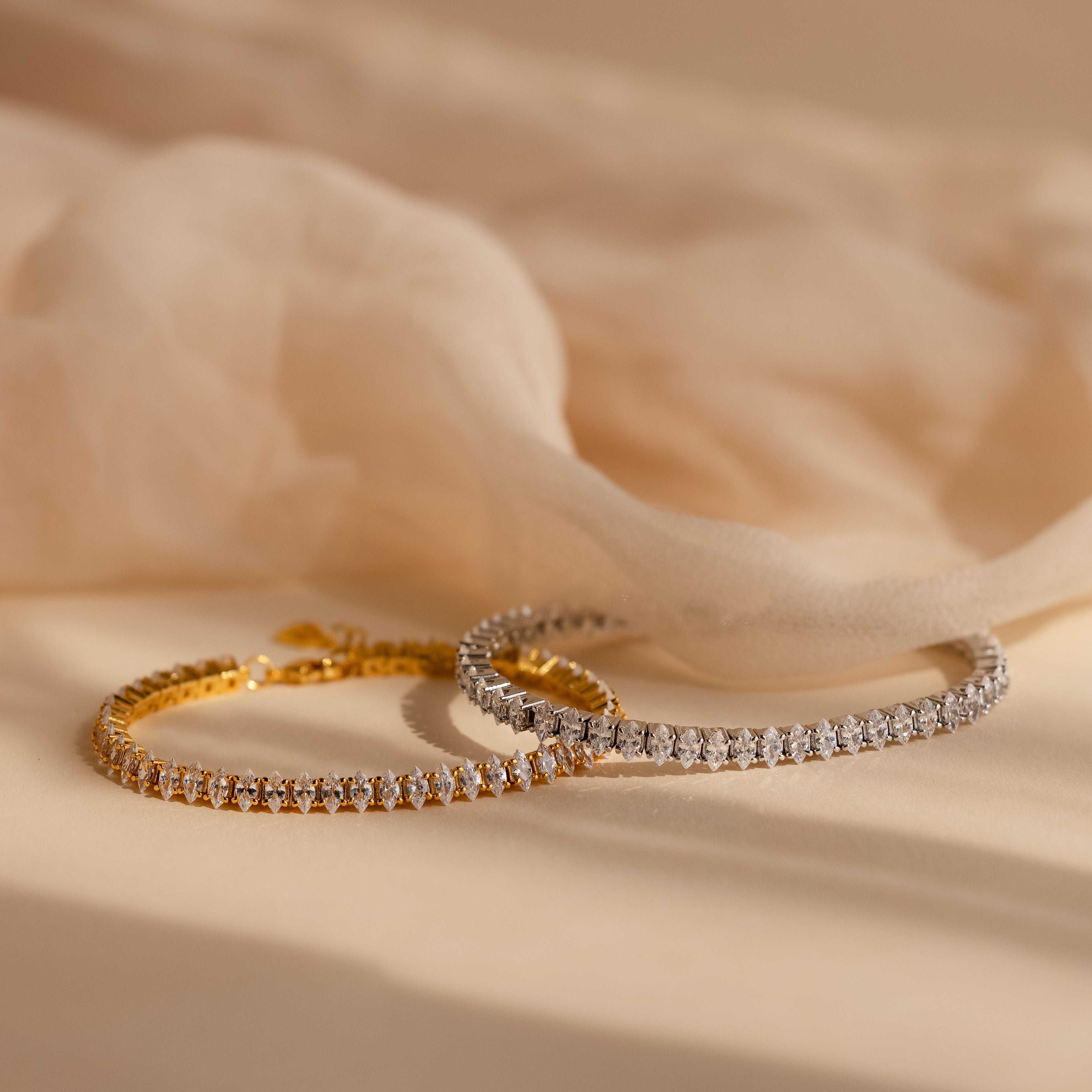 Side-by-side display of the Marquise Diamond Tennis Bracelet in silver and gold, both sparkling with marquise-cut diamonds, resting on soft fabric.