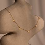The Heart Station Necklace in 18K Gold, featuring delicate heart charms on an elegant cable chain, is styled over a textured, abstract beige torso sculpture.