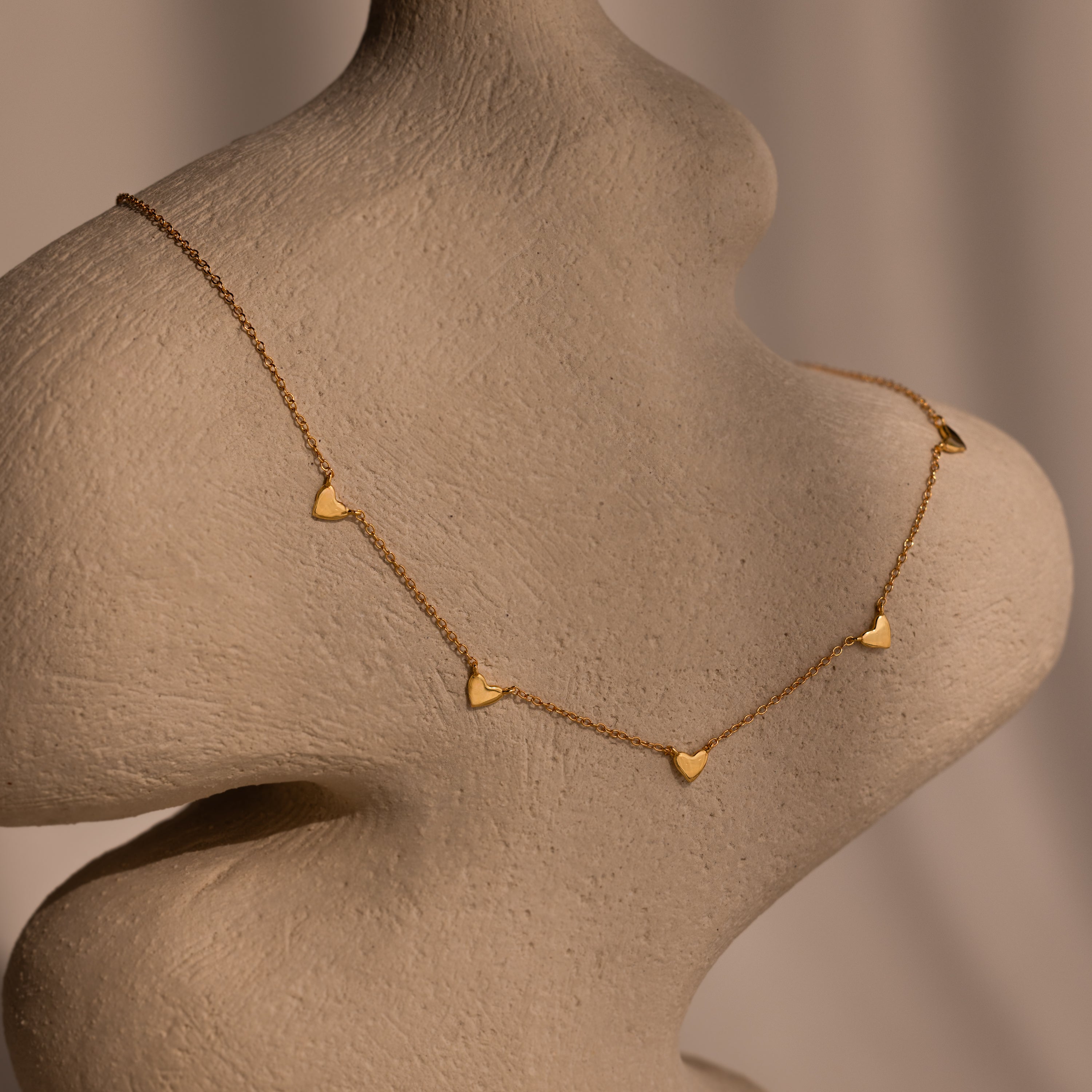 The Heart Station Necklace features delicate heart charms on a classic cable chain, showcased on a textured beige mannequin bust under soft lighting.