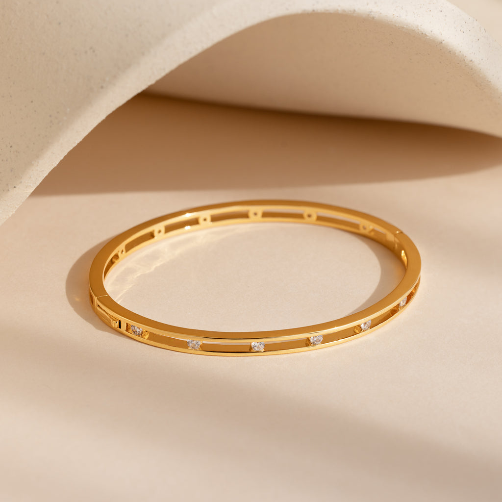 The Lace Diamond Bangle, crafted in gold and accented with delicate diamonds, is showcased on a beige surface with a curved backdrop—an ideal piece for luxury jewelry enthusiasts.