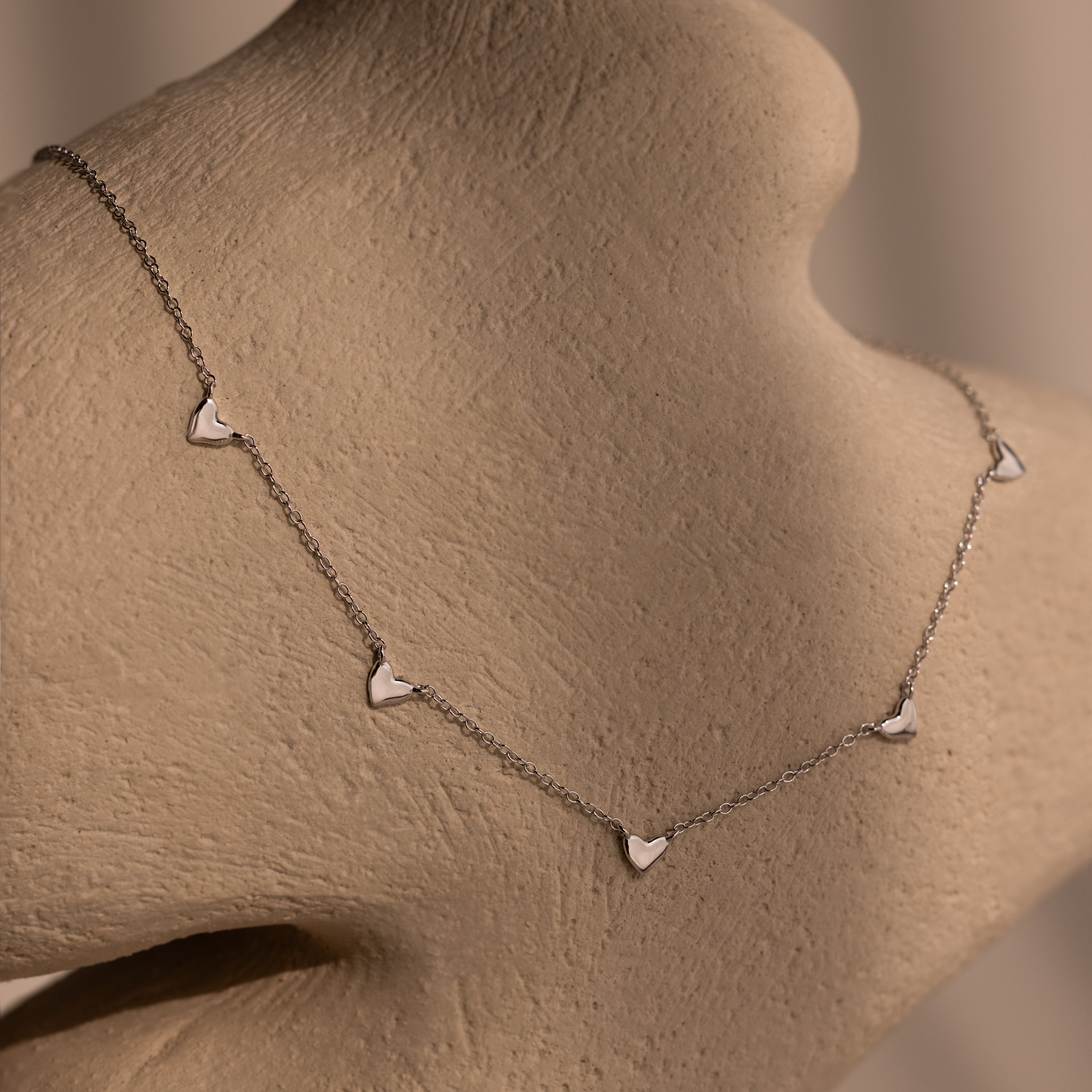 Heart Station Necklace in Silver, featuring delicate heart charms on a cable chain, displayed on a beige textured mannequin bust.