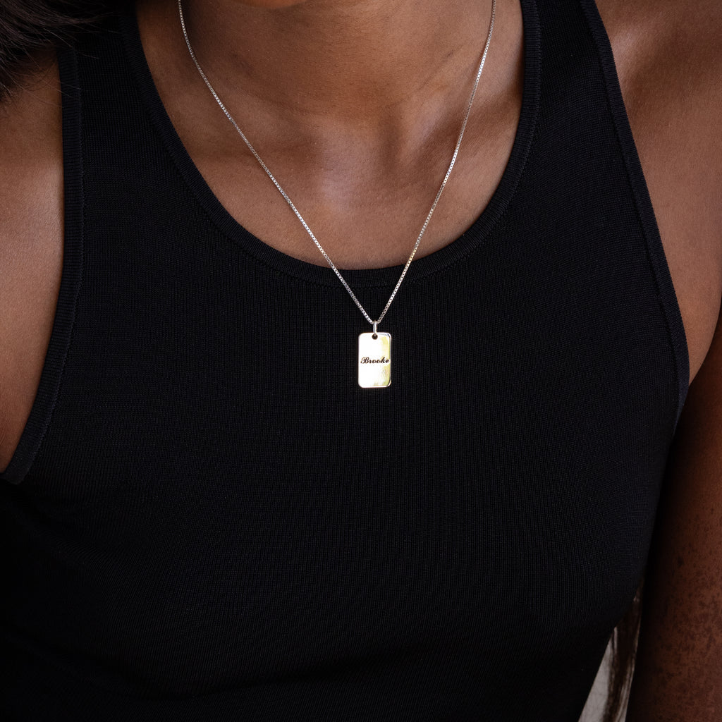 Woman in a black tank top wears the Name Tag Necklace in Sterling Silver, featuring a rectangular gold pendant for a modern, chic look.