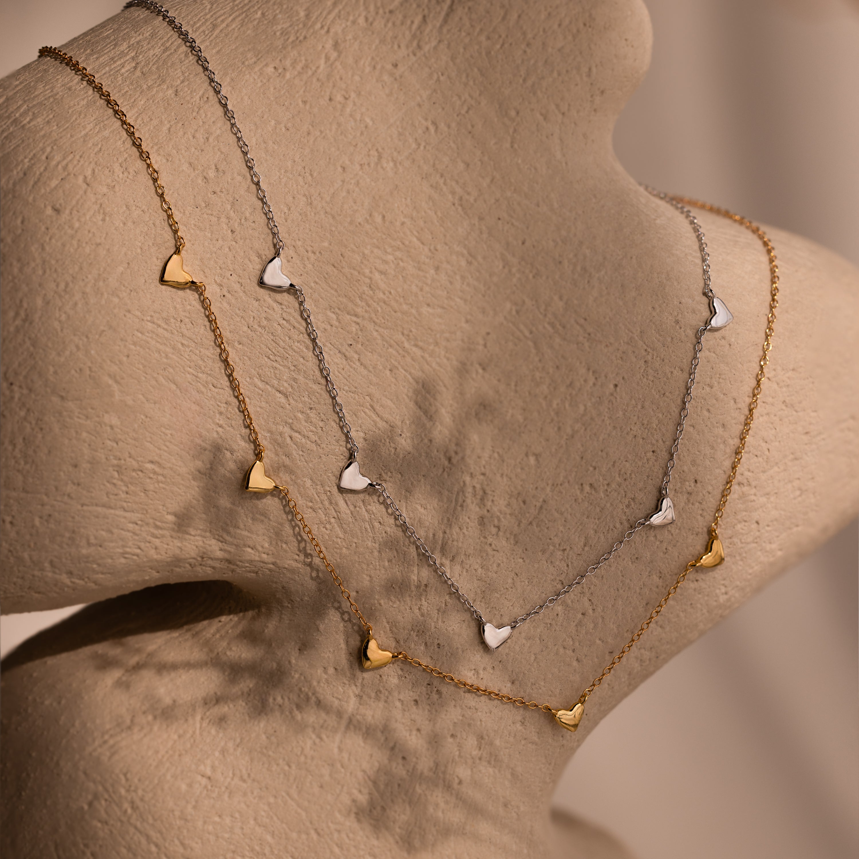 The Heart Station Necklace in 18K Gold features heart charms and is elegantly displayed on a neutral textured mannequin bust.