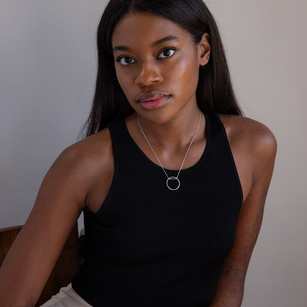 A woman with long dark hair, wearing a black sleeveless top and the Inner Circle Necklace in Sterling Silver—a striking silver circle pendant and chain—sits indoors.