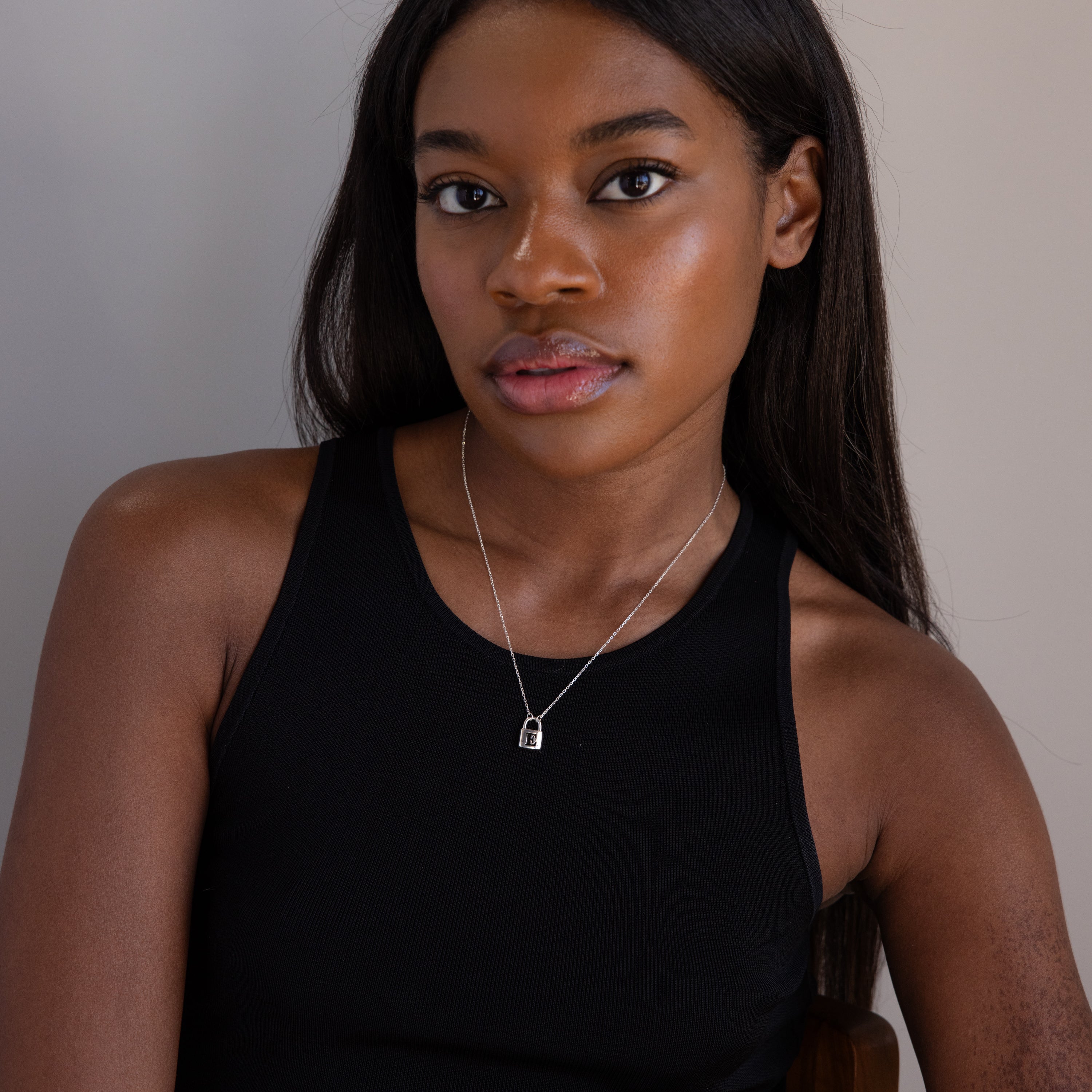 A woman wearing a black tank top models the Padlock Necklace in silver, sitting against a neutral backdrop and gazing into the camera for an understated fashion statement.