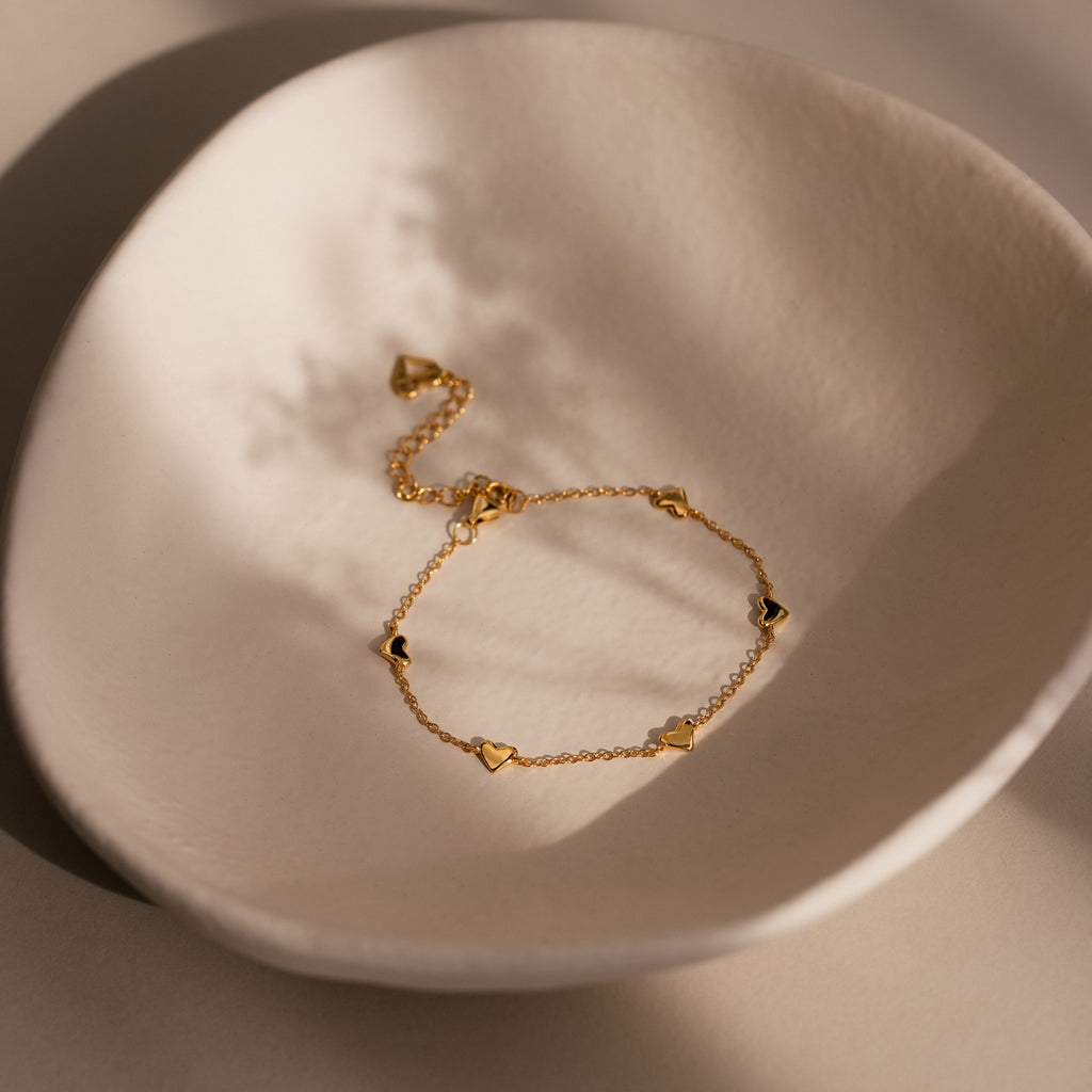 Heart Station Bracelet in 18K Gold, featuring delicate heart charms, is a romantic stacking piece shown on a textured off-white ceramic dish under soft light.